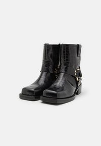 Botas de tobillo de charol negro con un patrón de cocodrilo, punta cuadrada y acentos de hardware dorado. Tacón bajo resistente y detalle de correa envolvente.