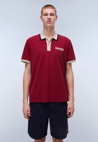 Polo bordeaux con colletto e bordature delle maniche beige, caratterizzato da un logo sul petto. Indossato con pantaloni corti blu scuro, che mostrano una vestibilità comoda.