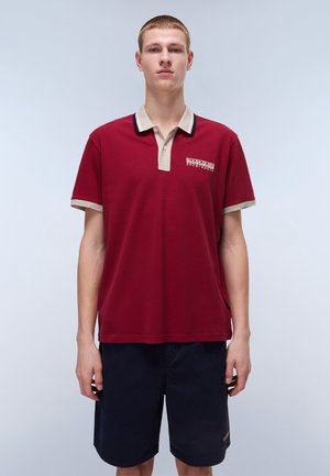 Bordeaux polo-shirt med beige krave og ærme trim, med et logo på brystet. Båret med mørkeblå shorts, der viser en afslappet pasform.