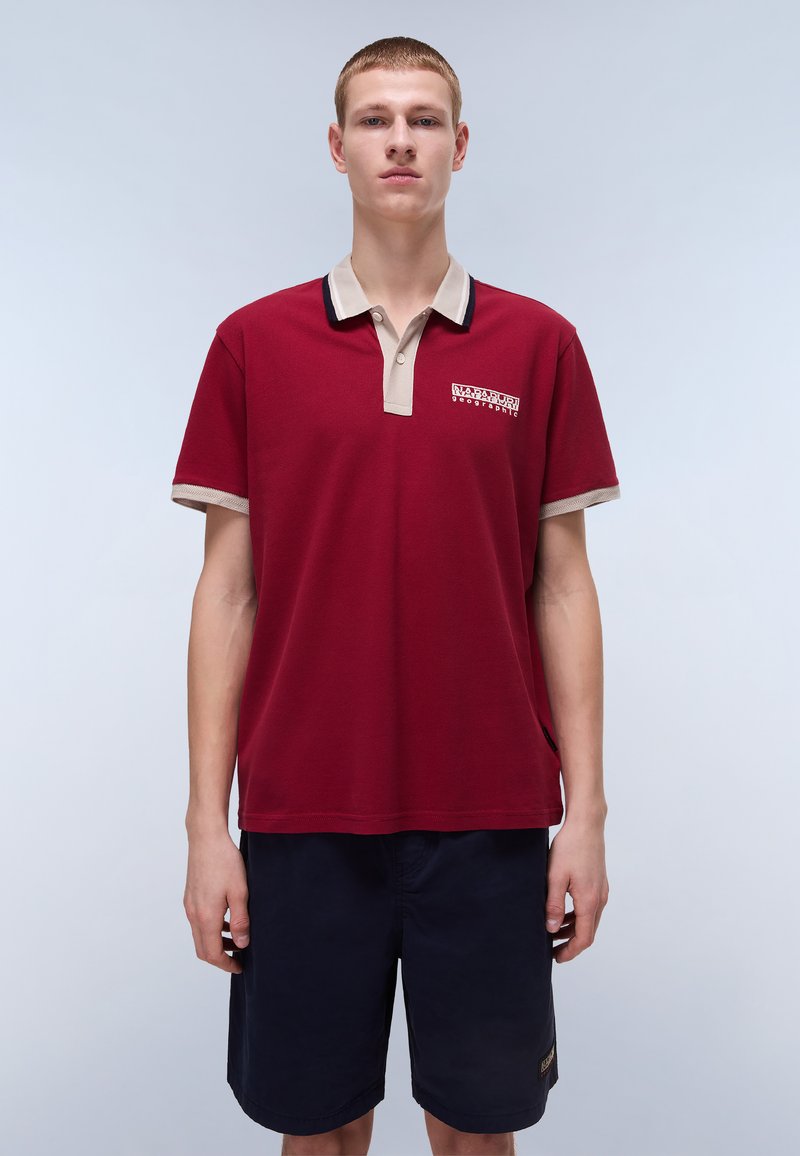 Polo bordeaux con colletto e bordature delle maniche beige, caratterizzato da un logo sul petto. Indossato con pantaloni corti blu scuro, che mostrano una vestibilità comoda.