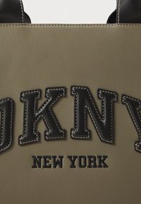 Alyvuogių spalvos maišas su juodais odiniais akcentais. Pasižymi iškilmingu tekstu "DKNY NEW YORK", ryškiais siūlų linkiais ir struktūrizuotu dizainu.