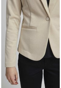 Beige texturerad kavaj med en enda knappstängning, som har två framfickor och en skräddarsydd design, bärs över en mörk topp och byxor.