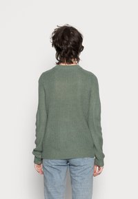 Olivgrüner Strickpullover mit gerippter Textur, langen Ärmeln und rundem Halsausschnitt, kombiniert mit hellen Jeans. Rückansicht gezeigt.