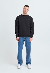 Schwarzer Pullover mit Rundhalsausschnitt und gerippten Bündchen, kombiniert mit hellblauen Jeans. Weiße Sneaker vervollständigen das Outfit. Schlichtes Design.