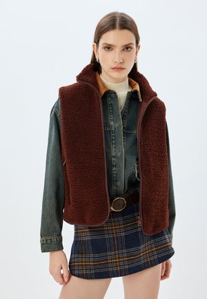 Bruine fleece vest met hoge kraag en ritssluiting, gelaagd over een denim shirt, gecombineerd met een geruite minirok in blauwe en gele tinten.