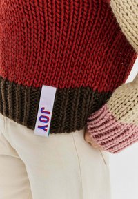 Sweter dziany z czerwonym korpusem, ciemnobrązowym obszyciem i wielokolorowymi rękawami. Metka z napisem "radość" w kolorowych literach.