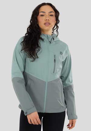 Donna con capelli lunghi e ricci che indossa una giacca outdoor bicolore grigio e verde acqua con cappuccio, tasche con zip e pantaloni neri.