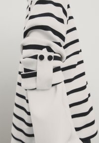 Poignet de pull à manches longues à rayures noires et blanches avec boutons noirs sur un vêtement à manches longues blanc uni.