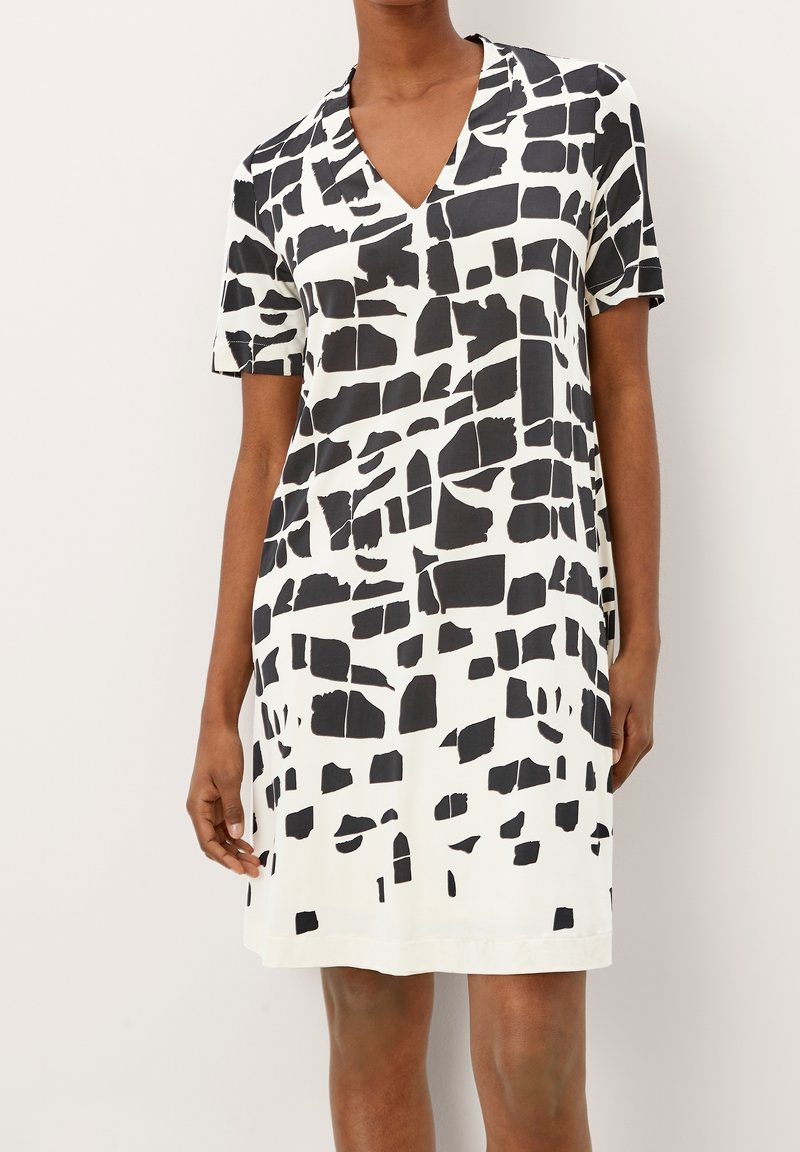 Femme portant une robe blanche à manches courtes, longueur genoux, avec un motif rectangulaire noir irrégulier et un col en V sur fond uni.