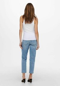 Witte tanktop, blauwe high-waisted jeans en zwarte heeled mules. Haar valt tot halverwege de rug; zichtbare stiksels op de jeans en achterzakken.