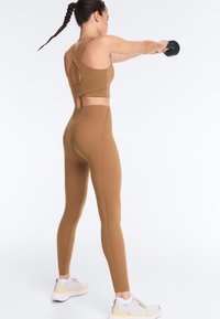 Femme en tenue de sport beige et baskets blanches effectuant un exercice de balancement avec kettlebell sur fond blanc uni.