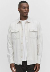 Lys beige skjorte lavet af blødt stof, med knaplukning, to brystlommer og lange ærmer, lagdelt over en hvid t-shirt.