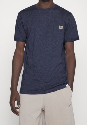 Mężczyzna noszący granatową koszulkę z krótkim rękawem z małą beżową łatą na klatce piersiowej oraz beżowe spodnie z małym logo na udzie.