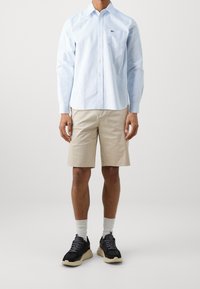 Jasnoniebieska, w paski koszula z guzikami, z kieszonką, w połączeniu z beżowymi szortami. Model ma na sobie czarne sneakersy i białe skarpetki; gładkie tło.