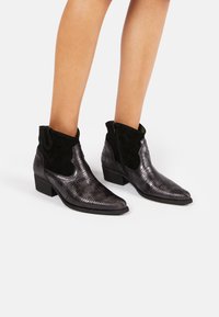 Schwarze Knöchelstiefel mit einem strukturierten, glänzenden Reptilmuster, Wildlederakzenten, spitzer Zehenform und einem niedrigen Blockabsatz. Seitenreißverschluss für ein einfaches An- und Ausziehen.