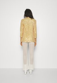 Blazer oro con paillettes sopra pantaloni a zampa d'elefante bianchi con motivo in pizzo, abbinato a scarpe con tacco alte bianche, visto da dietro.
