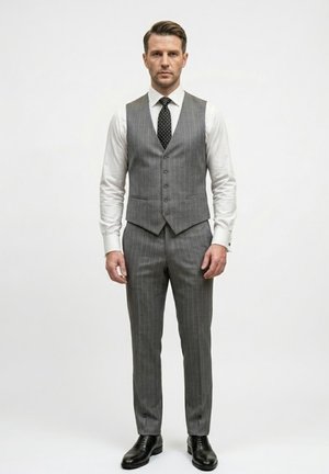 Hombre de pie contra un fondo liso, vestido con chaleco y pantalones gris rayados, camisa blanca, corbata negra con lunares blancos y zapatos de vestir negros.