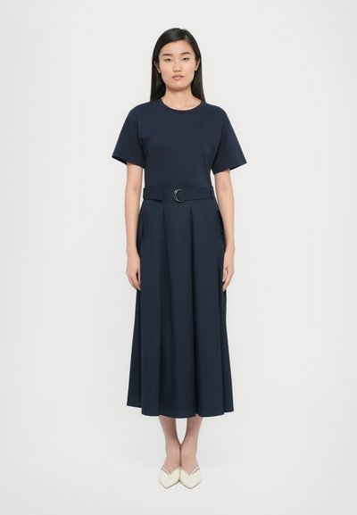 Marineblauwe t-shirt gecombineerd met een bijpassende lange rok met een ceintuurlijf en plooien. De outfit wordt aangevuld met crèmekleurige schoenen.