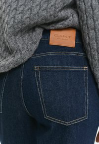 Mörkblå jeans med en läder GANT-etikett, fem fickor och kontrasterande sömmar. En grå stickad tröja täcker delvis midjebandet.