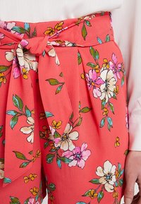 Pantaloni corta floreali corallo con vita annodata, realizzati in un tessuto testurizzato e decorati con vivaci motivi floreali in tonalità di rosa, giallo e verde.