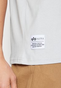 Lys grå bomulds t-shirt med struktureret stof, runde kanter og et rektangulært bomuldslabel med sort tekst og logo på siden.