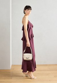 Bordeauxfarbenes Kleid mit Rüschelagen, knöchellang. Beige strukturierte Handtasche mit einem oberen Griff. Beigefarbene Riemchenheel runden den Look ab.