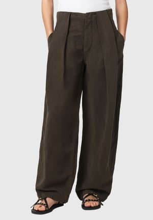 Personne portant un pantalon ample marron foncé à jambes larges et des sandales plates noires avec des œillets dorés, mains dans les poches.