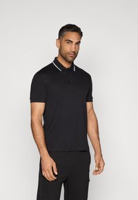 Černé polo tričko s límečkem, se bílým pruhem. Vyrobeno z hladkého materiálu, má krátké rukávy a vyšívané logo na hrudi.