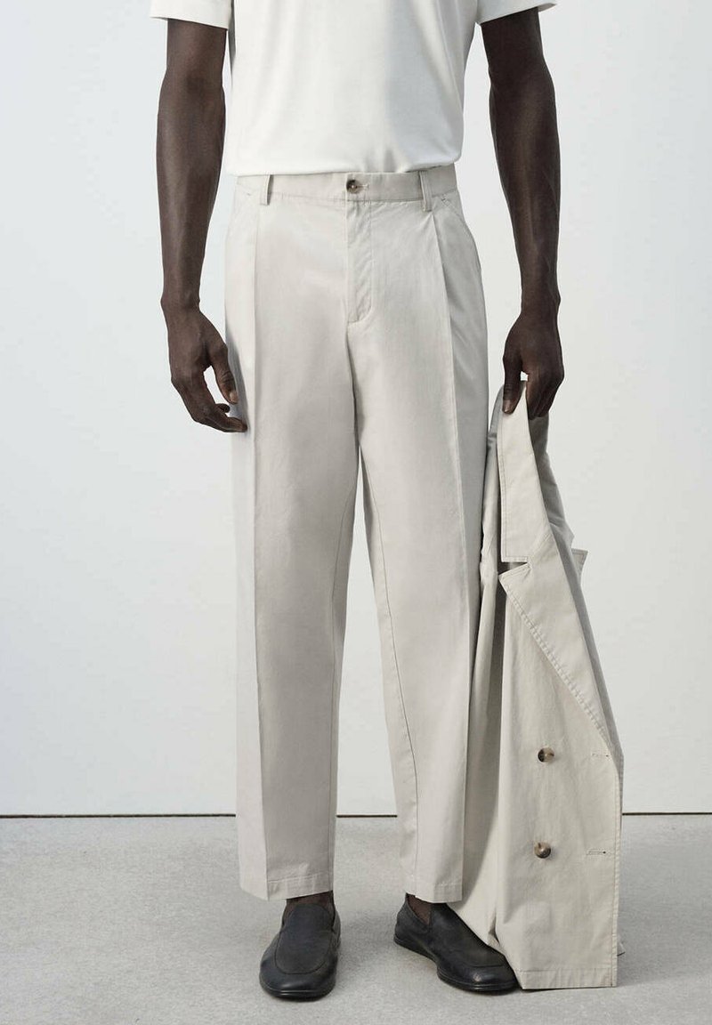 Homme portant un pantalon beige clair, une chemise blanche, des chaussures noires, tenant un manteau beige assorti dans sa main droite.