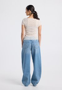 Nainen, jolla on tumma poninhäntä ja yllään valkoinen pitsinen lyhythihainen paita sekä väljen vaaleansiniset farkut, seisoo selin.