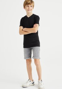 T-shirt nera in cotone con scollo a V, pantaloni corti di denim grigi con orlo arrotolato e sneakers bianche con dettagli blu navy. Il bambino è in piedi con le braccia incrociate.