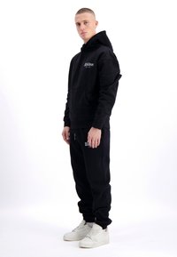 Zwarte hoodie met voorzak en logo, gecombineerd met zwarte joggingbroek met een merklogo. Model draagt witte sneakers.