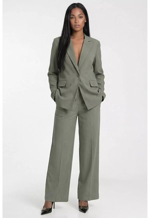 Kobieta stoi ubrana w oliwkowy garnitur w prążki z szerokimi nogawkami i marynarką oraz czarne, czubate wysokie obcasy, ręce w kieszeniach, na białym tle.