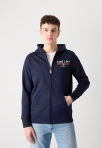 Veste à zip bleu marine avec une poche kangourou à l'avant, logo GANT sur la poitrine, tissu doux et poignets côtelés. Associée à un jean bleu clair.