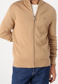 Beżowy sweter na zamek z miękkiego dzianinowego materiału z wysokim kołnierzem i ściągaczami na rękawach; posiada małe logo na lewej piersi, stylizowany z dżinsami.