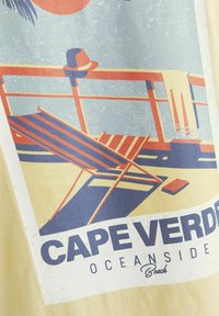 Grafisch ontwerp van een strandscène met gestreepte ligstoelen, een hoed en palmbomen, met de tekst 'Kaapverdië Aan Zee' op een beige achtergrond.