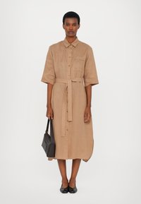 Beige, skjortekjole med belte i midjen, kort erm, brystlomme og knelangt fald. Kombinert med en svart håndveske og leiligheter.