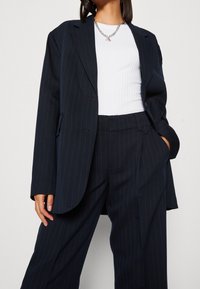 Abito a righe navy con una vestibilità comoda, composto da un blazer doppiopetto e pantaloni a gamba larga. Accompagnato da un top bianco a coste.