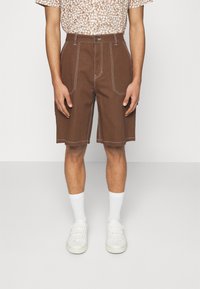 Shorts de algodón marrón con costuras blancas, que presentan una cintura alta y piernas anchas, combinados con calcetines blancos y zapatos slip-on blancos.