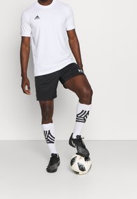 Vit idrottströja med upprullade ärmar, svarta shorts, mönstrade svart-vita strumpor och svarta fotbollsskor, stående på en fotboll.