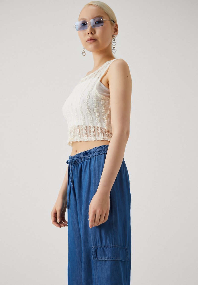 Un crop top en dentelle blanche avec une bande inférieure transparente, associé à un pantalon bleu ample à cordon de serrage et poches latérales. Lunettes de soleil hautes et boucles d'oreilles en cœur.