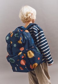 Mochila de color azul marino con patrones celestes coloridos (planetas, estrellas) y correas ajustables. Tejido suave con un bolsillo frontal; decorada con rayas.