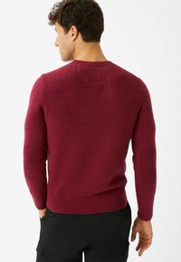 Roter Pullover mit geripptem Muster an den Schultern und Ellenbogen, taillierte Passform und Rundhalsausschnitt. Hergestellt aus weichem Strickmaterial.