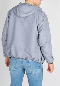 Grå hættejakke med elastisk bund og gule detaljer. Blødt materiale, der har en afslappet pasform og sidelommer. Kombineret med lyseblå jeans.