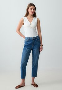 Top bianco a coste senza maniche con bottoni dorati, abbinato a jeans in denim blu a gamba dritta e sandali scivolati marroni. Abbigliamento semplice e casual.