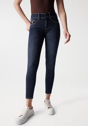 Jeans azul oscuro de corte ajustado combinados con zapatillas blancas, mostrando vista frontal desde la cintura hasta los tobillos sobre un fondo liso.