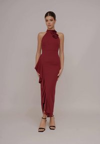 Woman wearing a sleeveless burgundy halter dress with ruffle detail and black ankle-strap heels, standing against a plain light background.