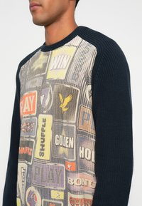 Maglione lavorato a maglia con un patchwork colorato di etichette grafiche; parte anteriore multicolore con un retro e maniche a coste navy scuro texturizzati.
