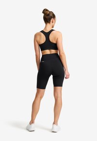 Kvinna med håret i en knut, iklädd svart sportbh, svarta högmidjade cykelshorts och vita sneakers, stående vänd bort mot en vit bakgrund.