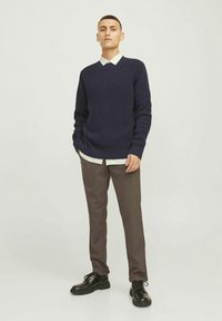 Marineblå strikkegenser med V-hals, lagd over en hvit skjorte med krage. Brune bukser og svarte snøresko fullfører antrekket.